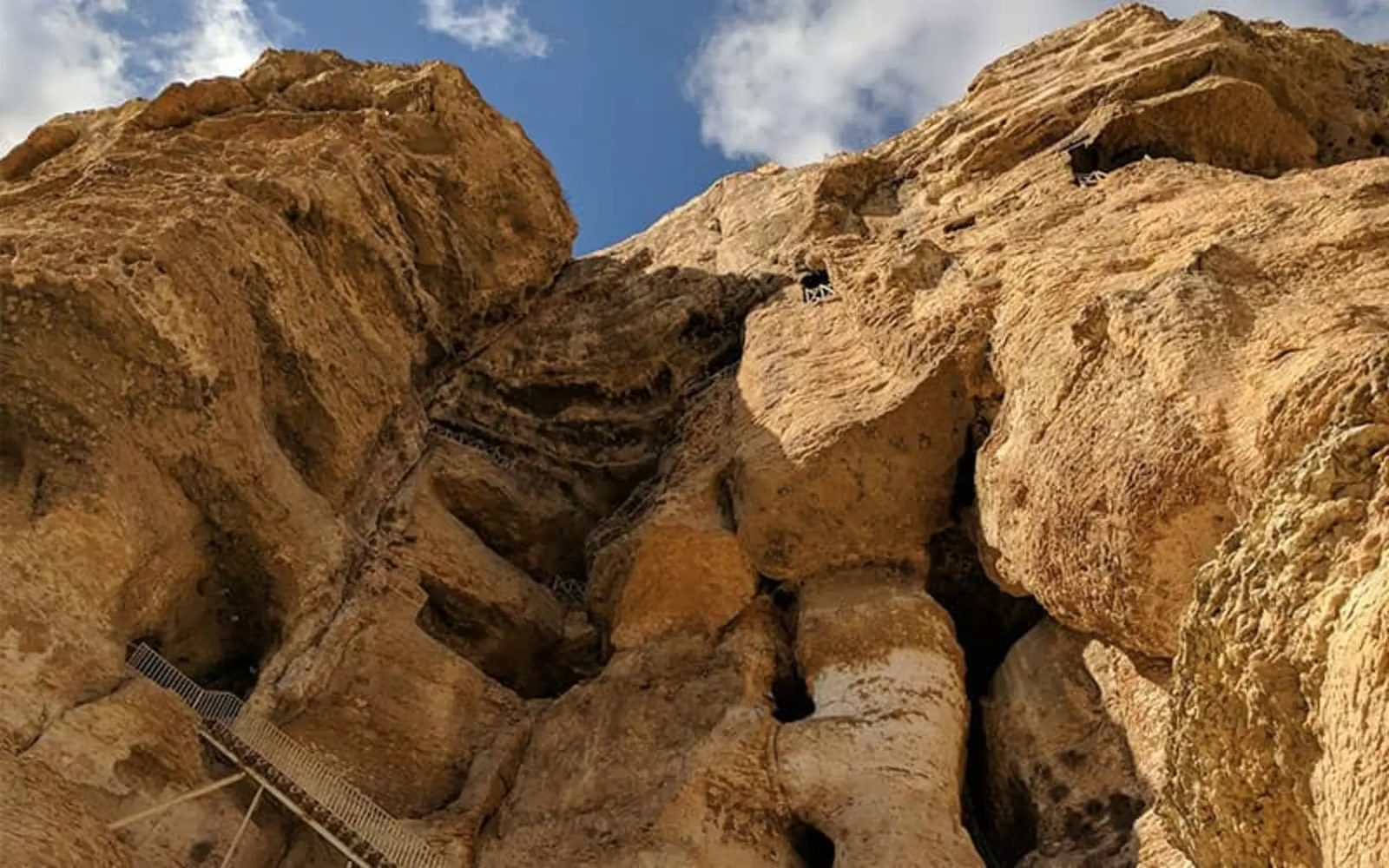 Cave openings high on the ochre limestone cliff of Karaftu Cave, with a metal stairway climbing toward them