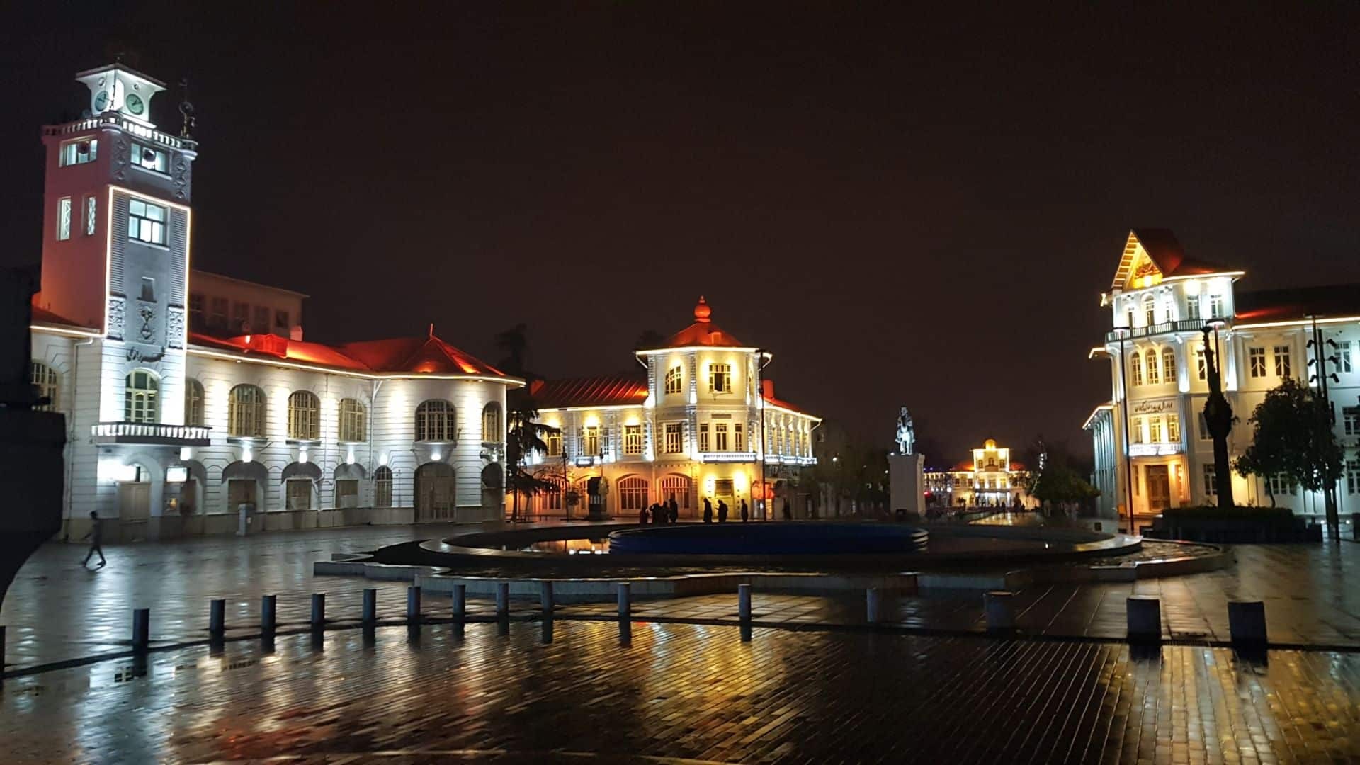 Qajar Kolah Farangi mansion lit at dusk across Sabzeh Square, with the equestrian statue of Mirza Kuchak Khan